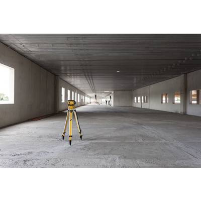 A surveying instrument stands in a large, empty, unfinished room with a concrete floor and high ceilings. Workers can be seen in the background.