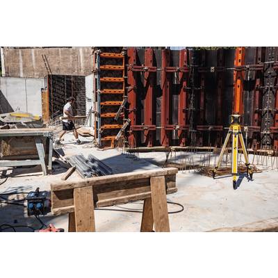 Construction site with a man assembling a wooden frame. Building tools and materials in the foreground. Wooden beams and measuring equipment are visible.