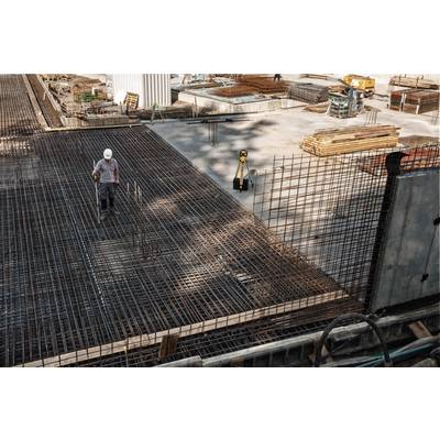 A worker wearing a hard hat walks across a construction site with steel reinforcements. Materials and machinery are visible in the background.
