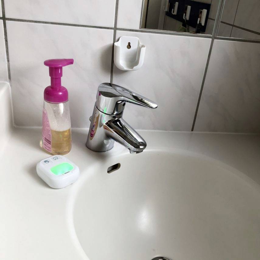 A bathroom sink with a silver tap. Beside it stands a pink soap dispenser and a small electronic device with a glowing green display.