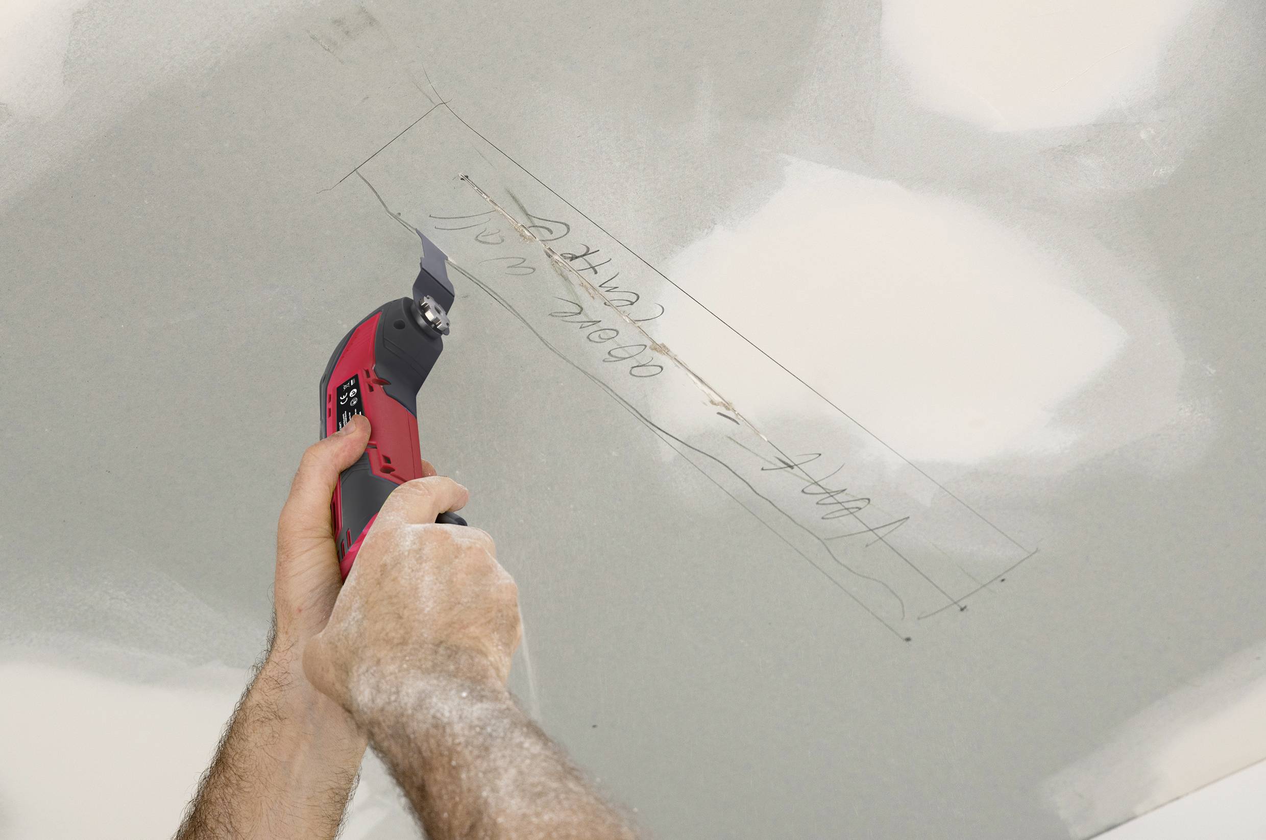 A person is cutting a rectangle out of plasterboard using a red electric saw. Markings can be seen on the board.
