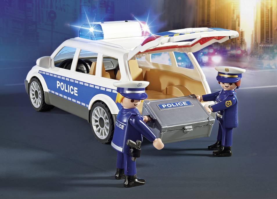 Two toy police officers load a suitcase marked 'Police' into the boot of a police car. The car has flashing lights.