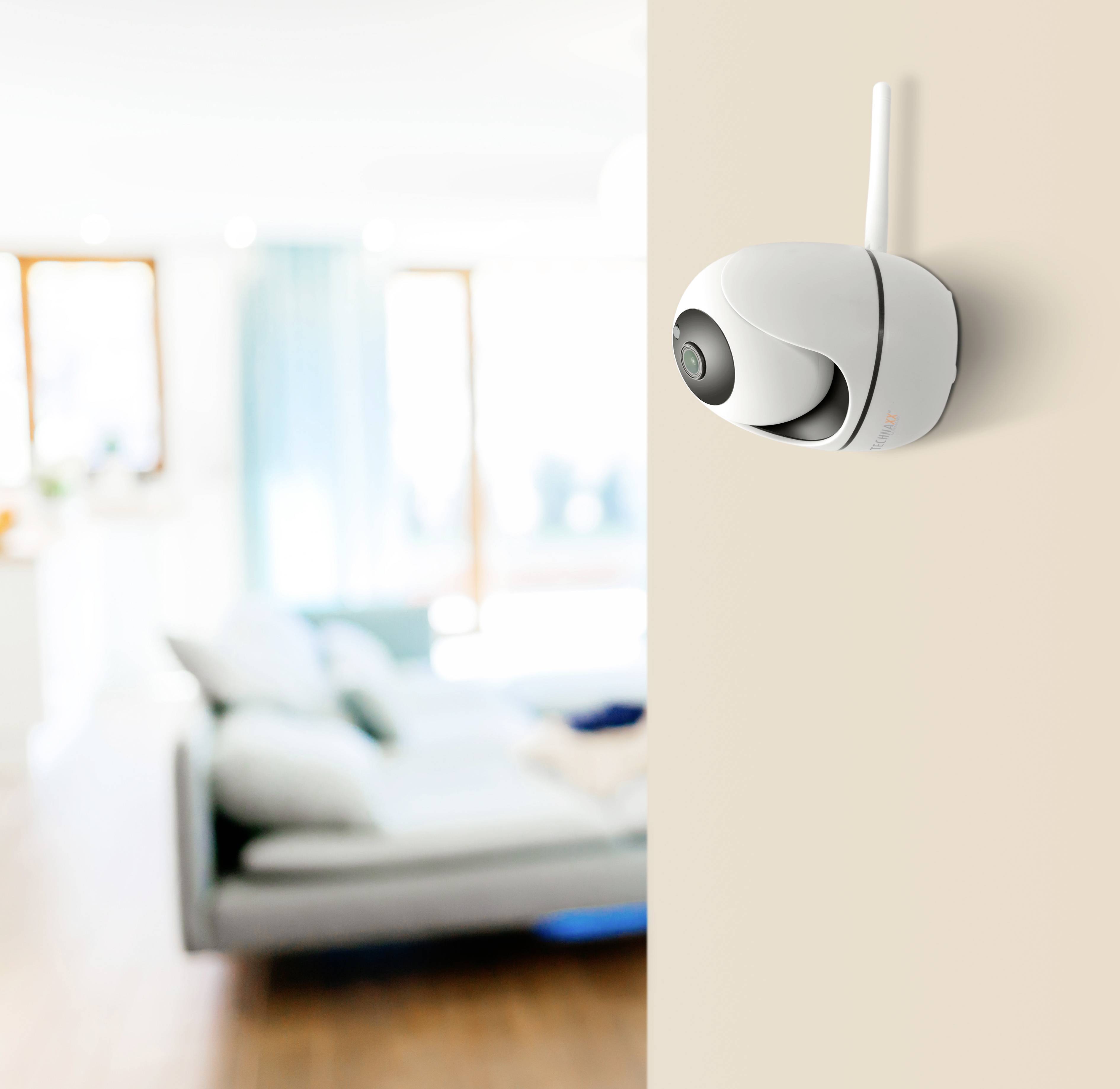 Security camera mounted on a wall, with a blurred living room and sofa in the background.