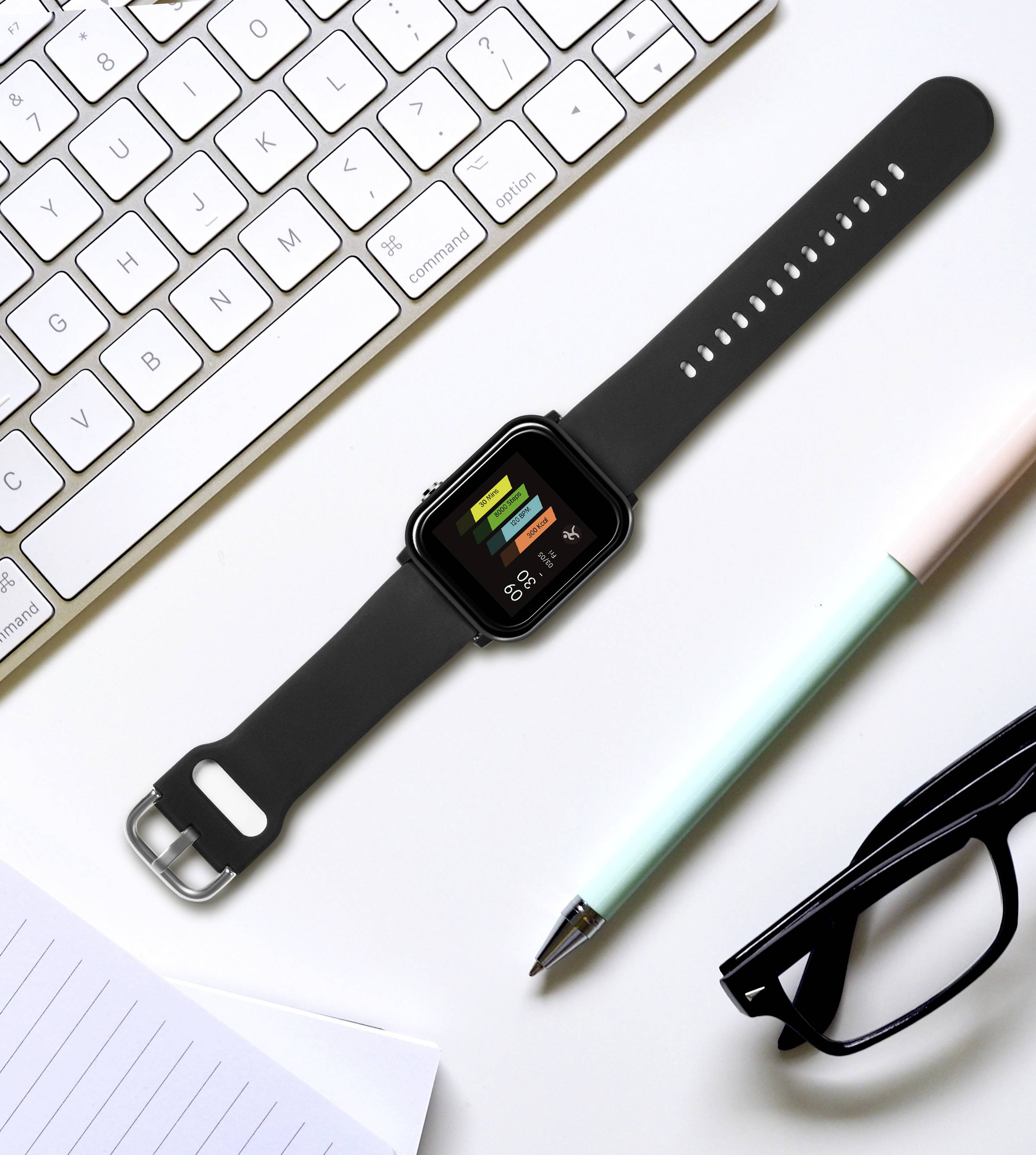 A watch with fitness display and colourful bars lies on a desk next to a keyboard, glasses, pen and notepad.