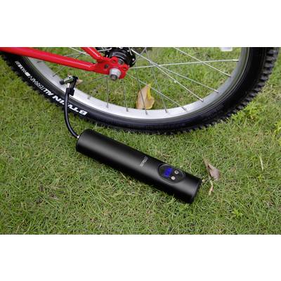 Black portable compressor inflating a bicycle tyre on green grass. Digital display on the compressor shows the pressure.