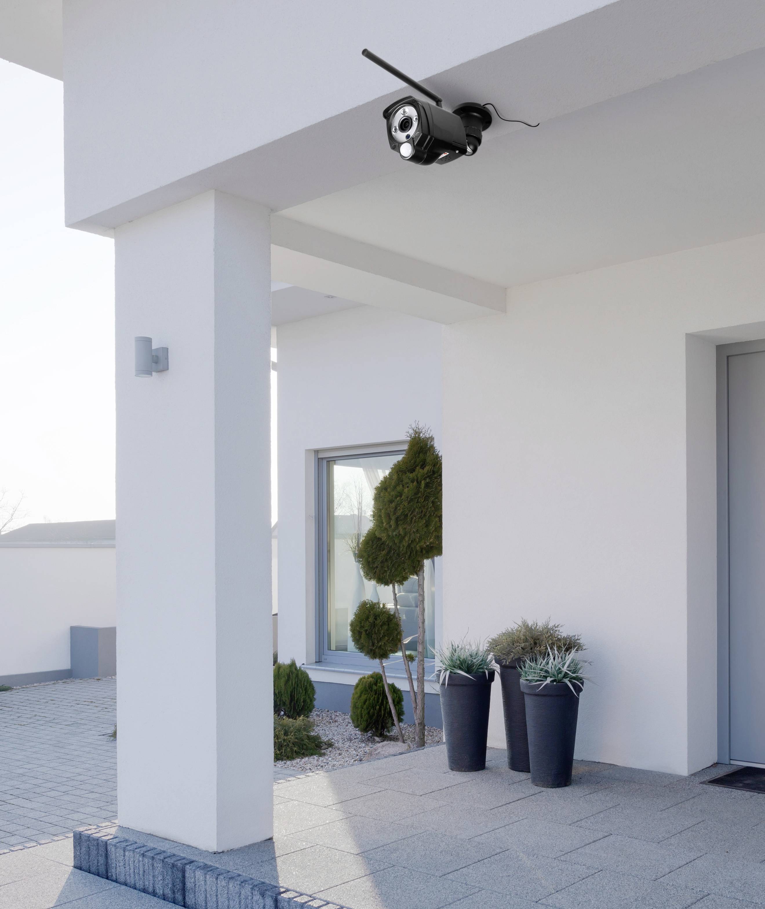 Security camera mounted on a modern house facade, monitoring the entrance area. Two potted plants stand beside the door.