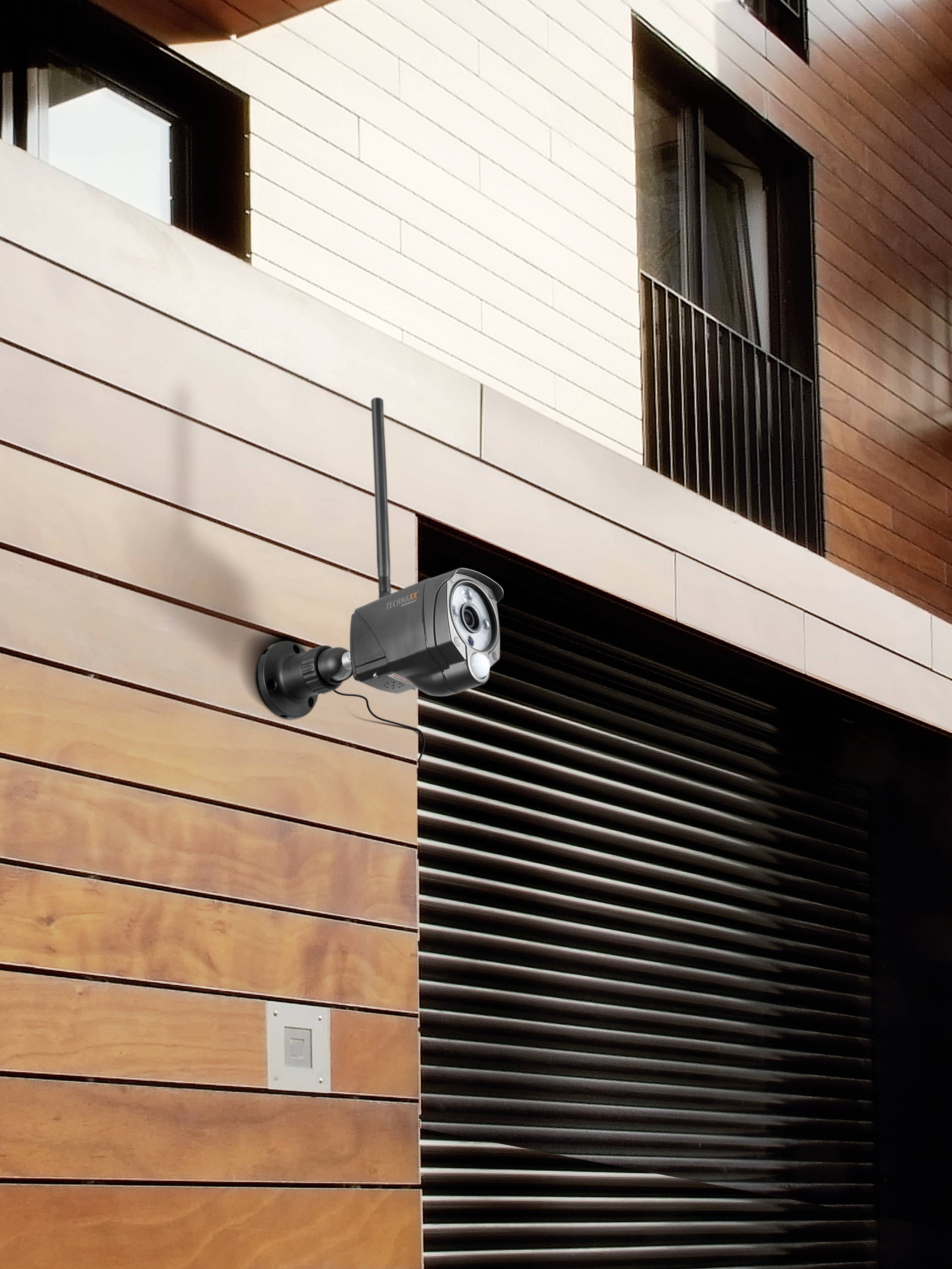 A security camera is mounted on a modern building with wooden elements, monitoring an area.