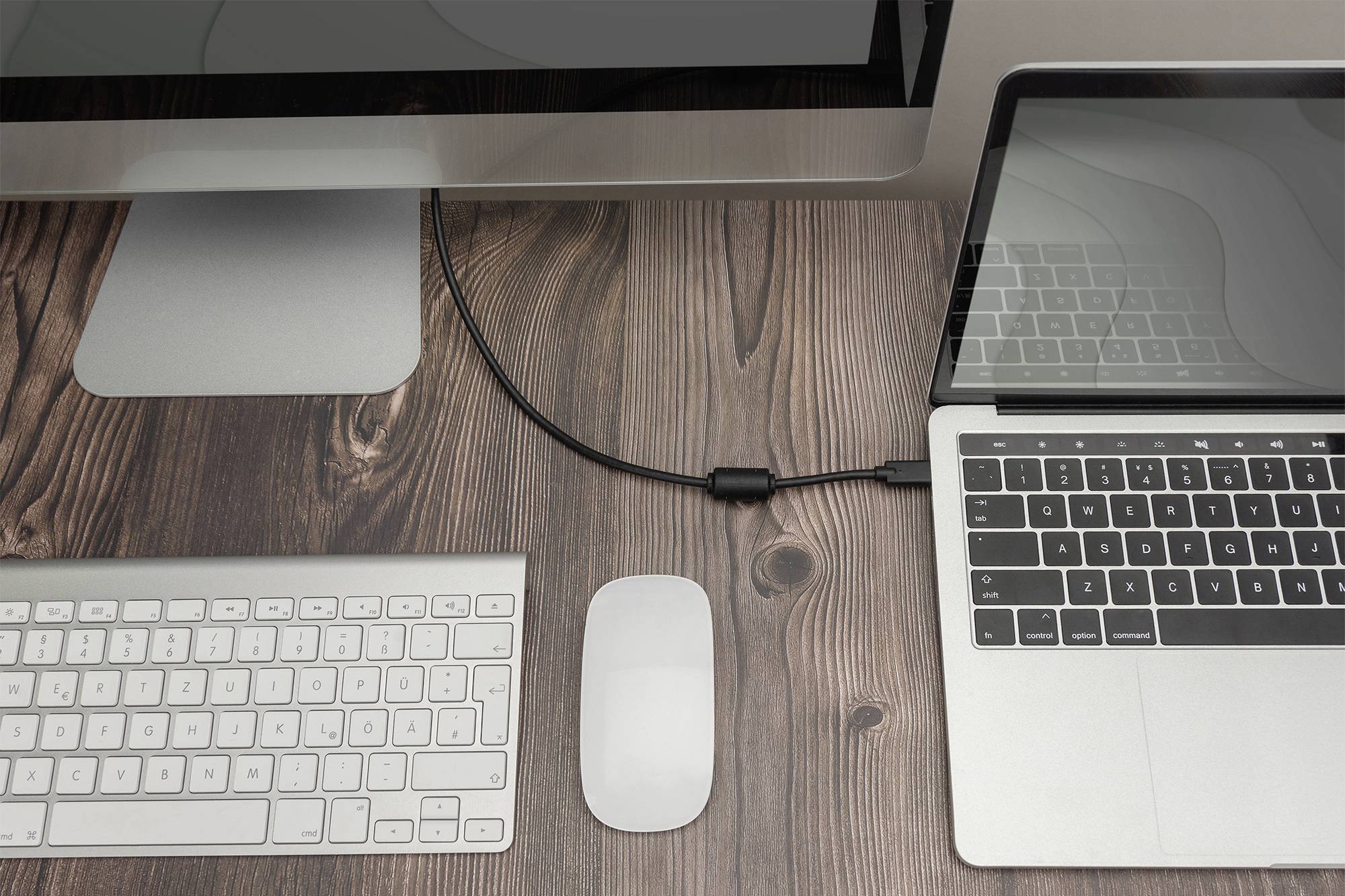 A laptop and a desktop computer are connected on a wooden table. A keyboard and mouse are lying beside them.