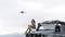 A woman is sitting on an off-road vehicle holding a mug, with a drone hovering in the air. The background is cloudy.