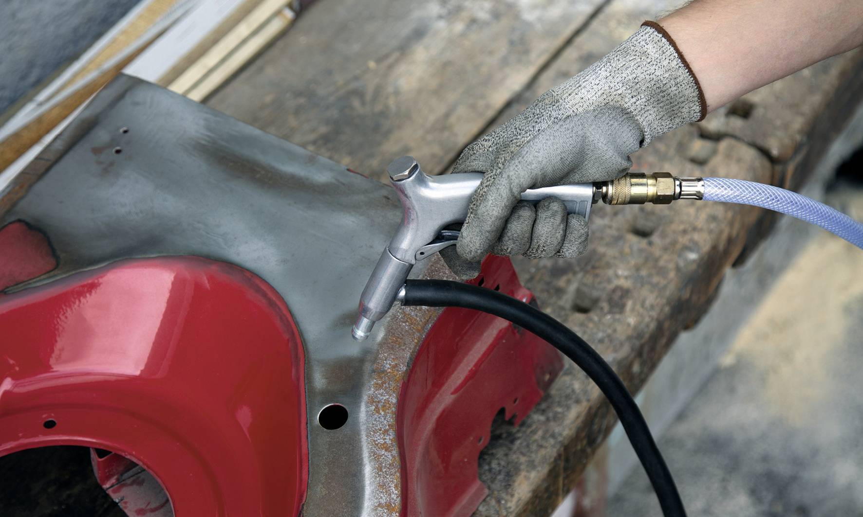 A person wearing grey work gloves is sandblasting a metal piece on a wooden surface; the paint is partially removed.