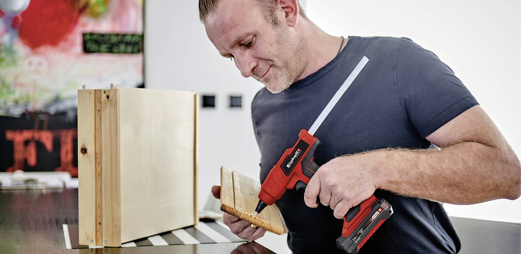 A man is securing a wooden board to a box using a cordless drill. A colourful picture is blurrily visible in the background.