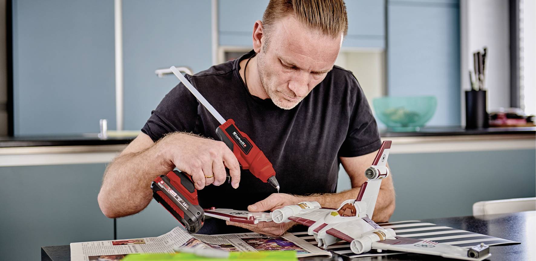 A man is working attentively with a cordless drill on a model aeroplane at a table in a modern kitchen.