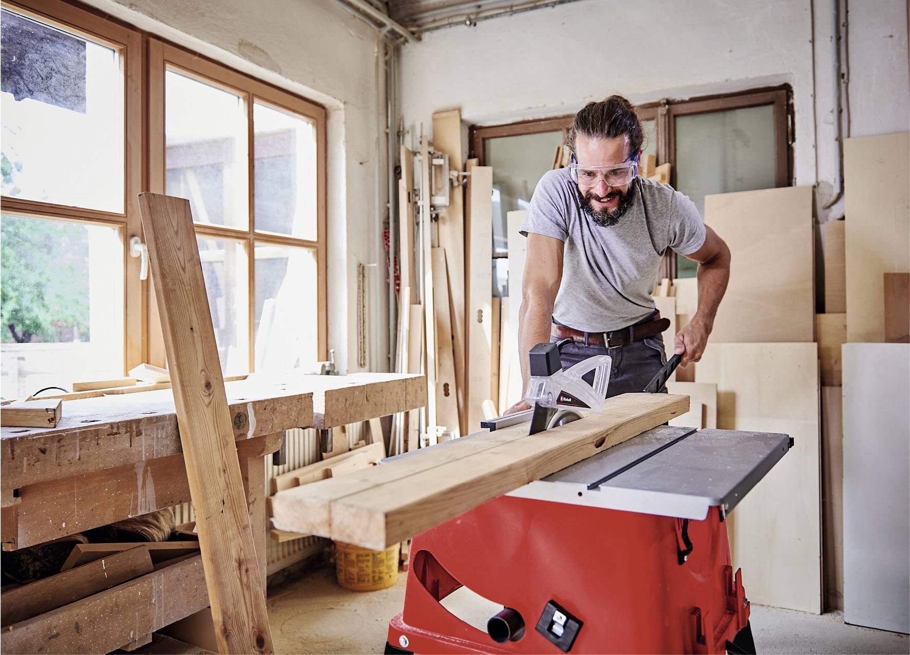 A man is working in a woodwork workshop, sawing a plank with an electric table saw. Surrounded by wood and tools.