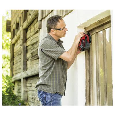 A man wearing glasses uses a handheld device to inspect a wooden window frame of a rustic building, focusing on the top corner.