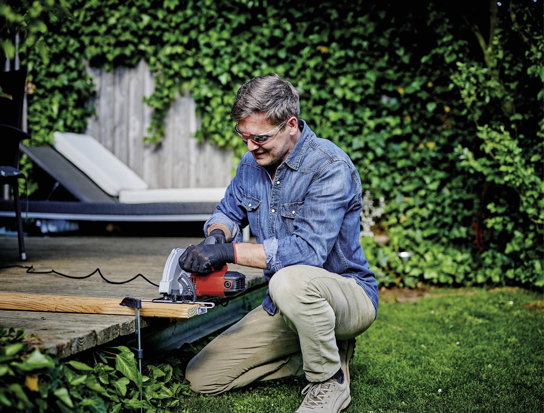 A man in work attire is sawing a wooden board with an electric saw in the garden. Plants can be seen in the background.