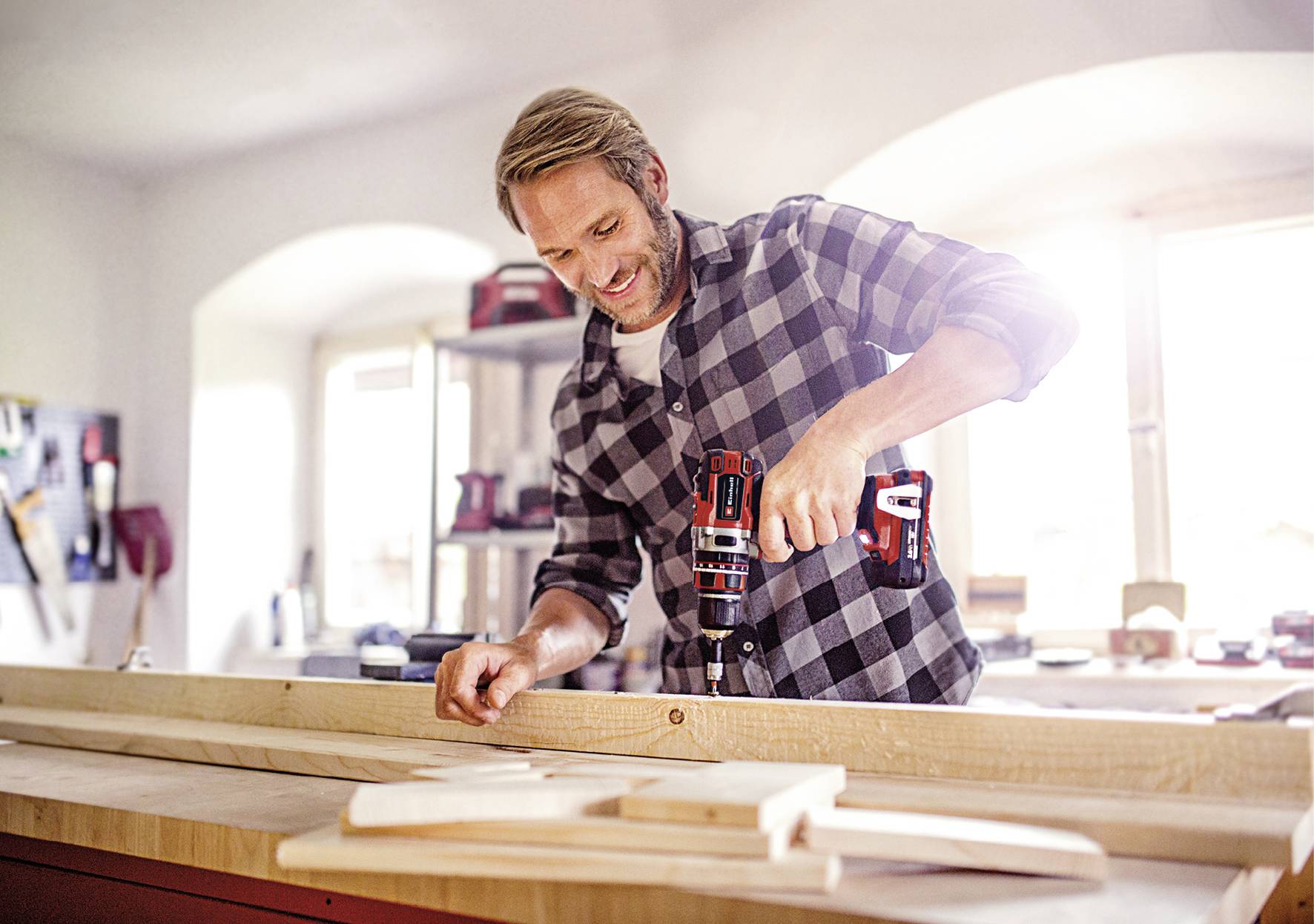 A man is working in a workshop and using a drill to work on wood at a workbench. He is smiling. Tools and windows can be seen in the background.