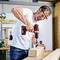 A man wearing safety glasses and a white T-shirt is drilling a hole in a wooden board using a cordless drill in a workshop setting.