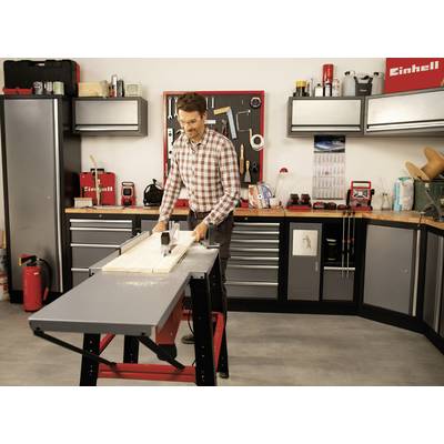 A man is working with a circular table saw in a workshop. Surrounded by tools and shelves, he is cutting wood to size.