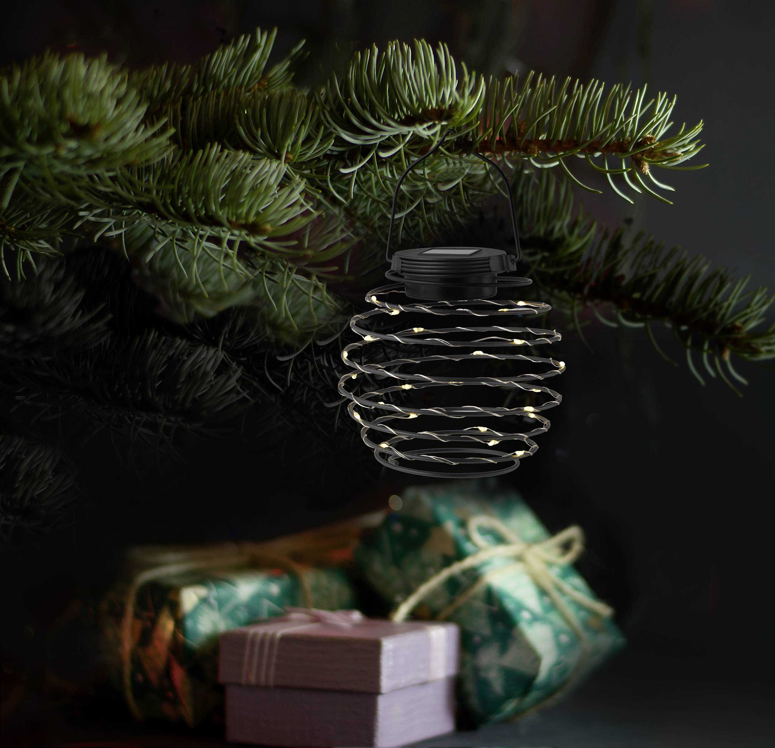 Lantern with fairy lights hangs on pine branches, with gifts wrapped in green and pink paper underneath. Festive Christmas decoration.