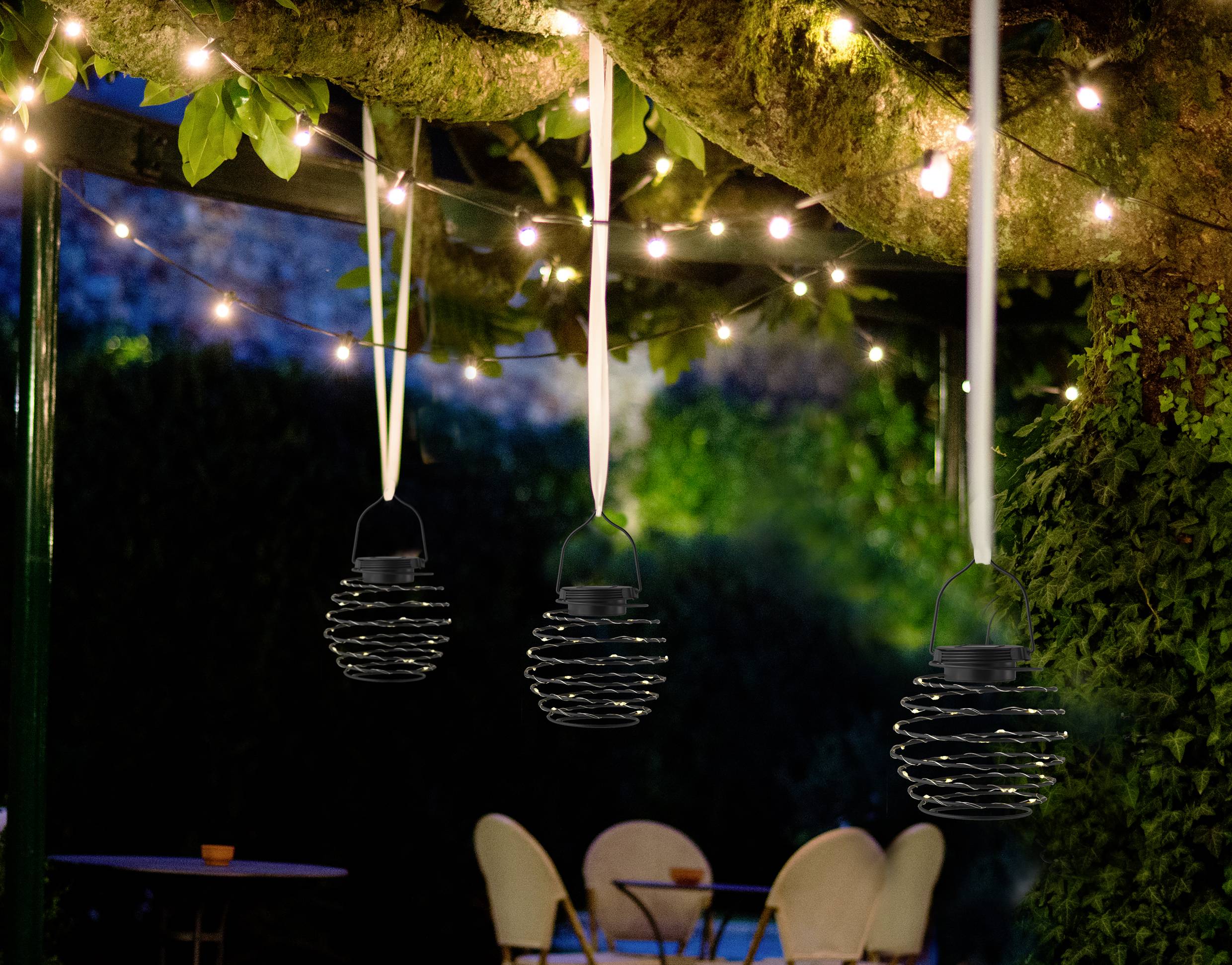 Three hanging lanterns beneath an illuminated tree in the garden. In the background, blurred chairs and a table are visible.
