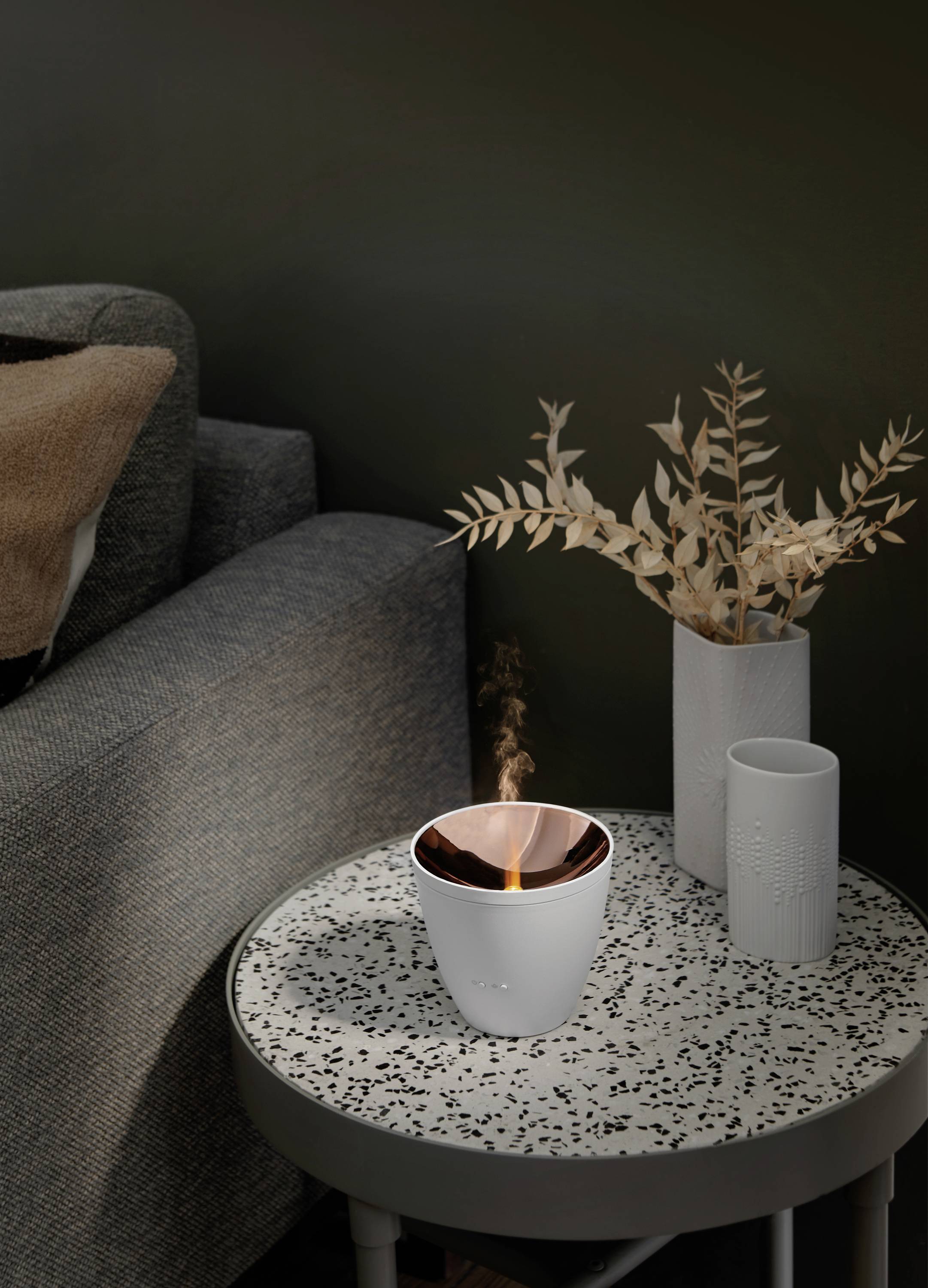 White candle in a modern container with a flame on a patterned table next to a grey sofa and a vase with dried branches in a stylish setting.