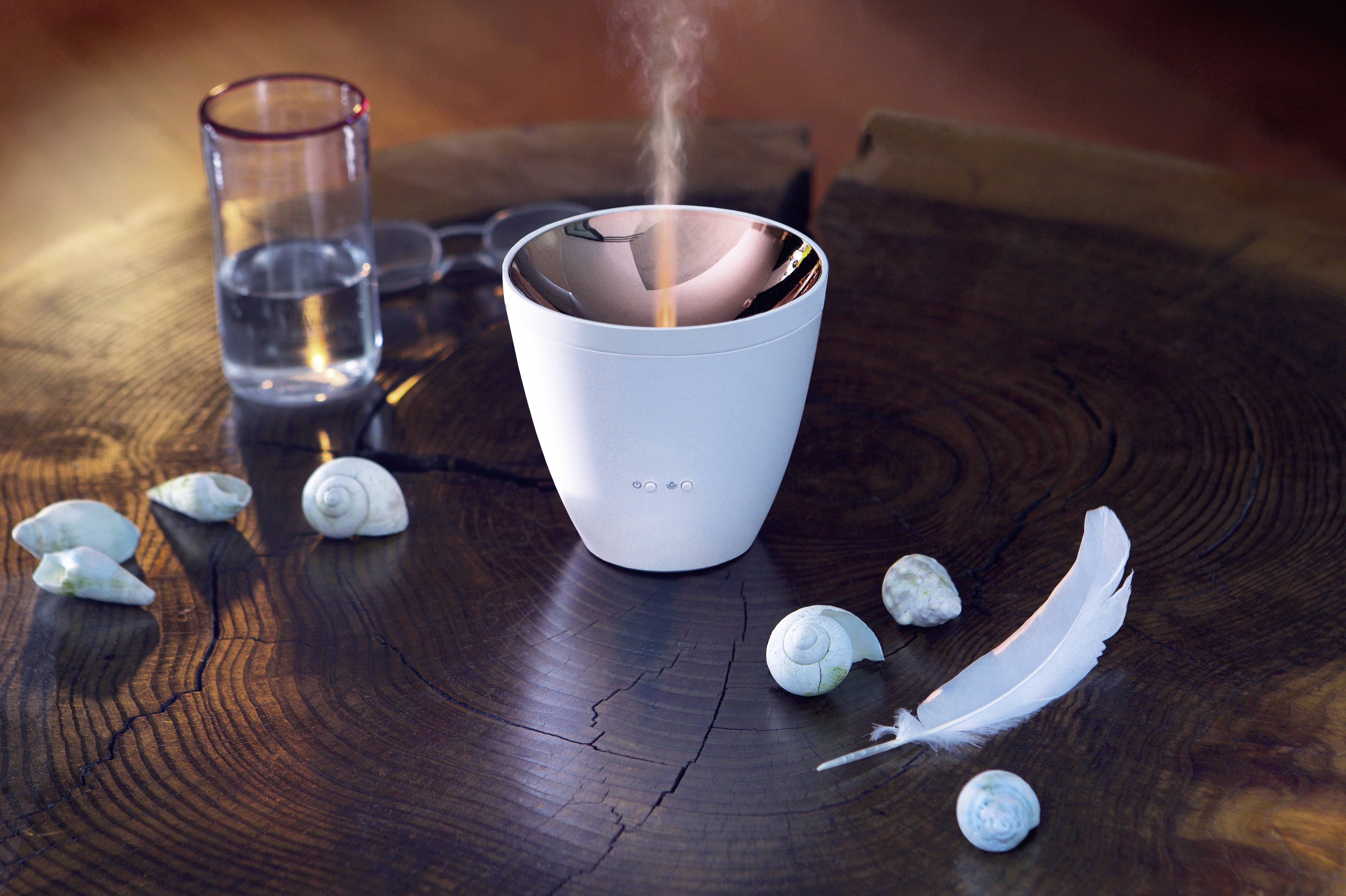 A white air humidifier on a wooden table, surrounded by seashells and a feather. A glass of water sits beside it.