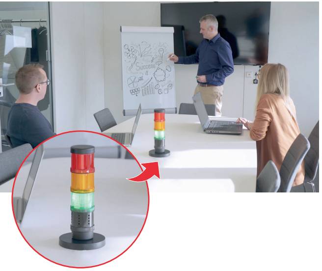 A man is presenting diagrams on a flipchart in a conference room. Two people are seated at the table, one with a laptop.