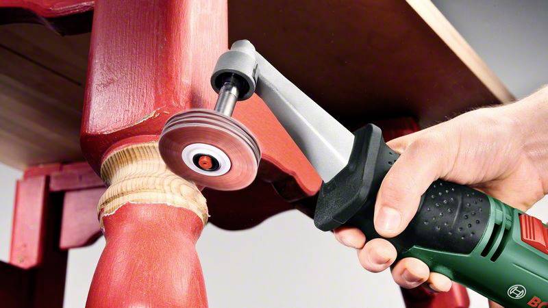 A hand holds a rotating grinding tool working on a red wooden table leg to remove paint.