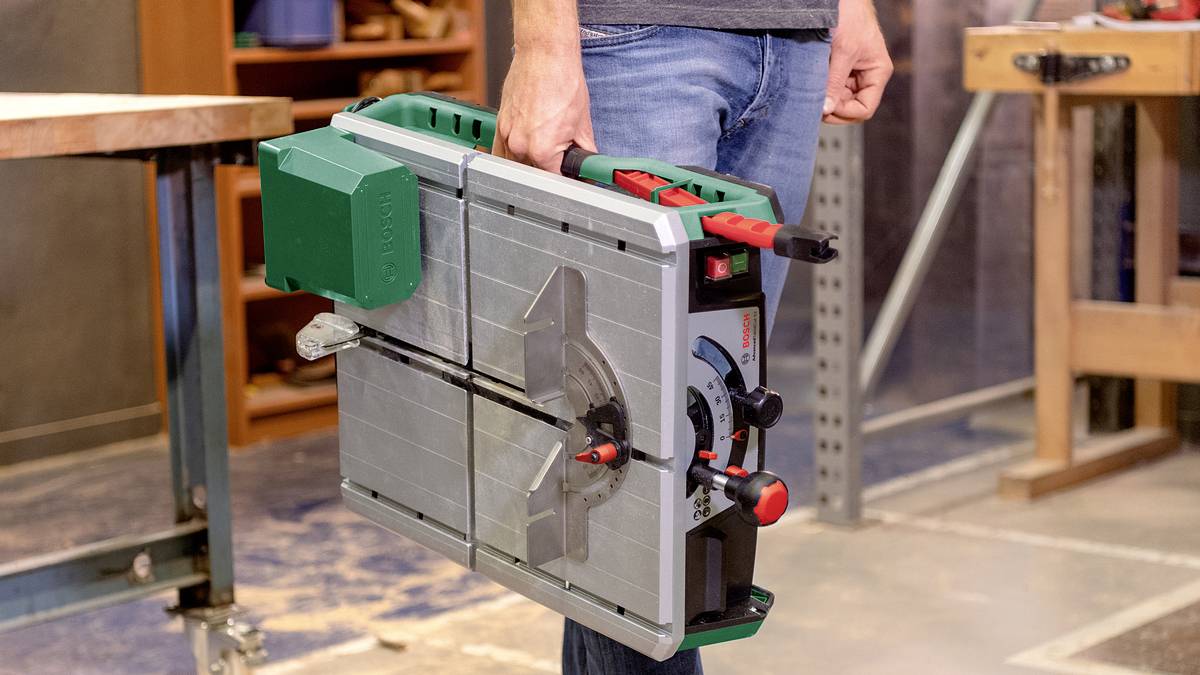 A person is holding a portable table circular saw in a workshop. The saw has a green guard and features red control elements.