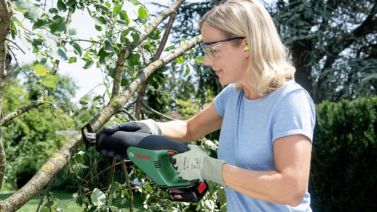 A woman wearing safety glasses and gloves is cutting branches in the garden with an electric saw.