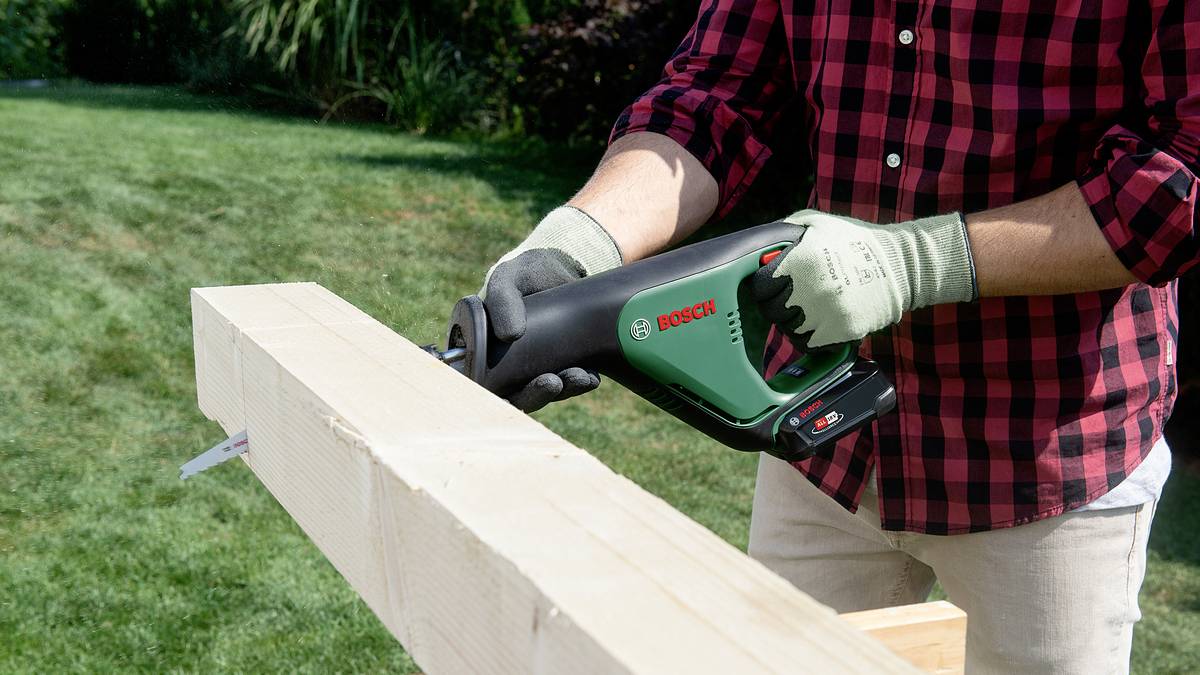 A person is sawing a wooden board in the garden using an electric reciprocating saw. They are wearing green gloves and a red-checked shirt.
