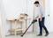 A man is standing in a workshop, using a cordless vacuum cleaner to clear sawdust from the floor. A workbench is visible in the background.