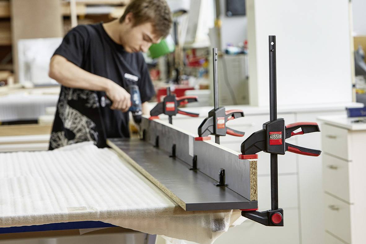 A man is working in a workshop and securing a board with clamps while drilling it.