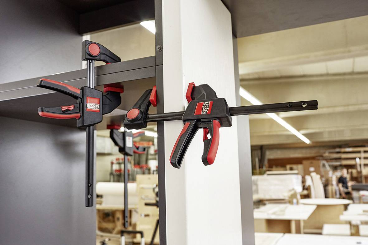Two red G-clamps are securing a white board to a grey shelf in a workshop. Woodworking items are visible in the background.