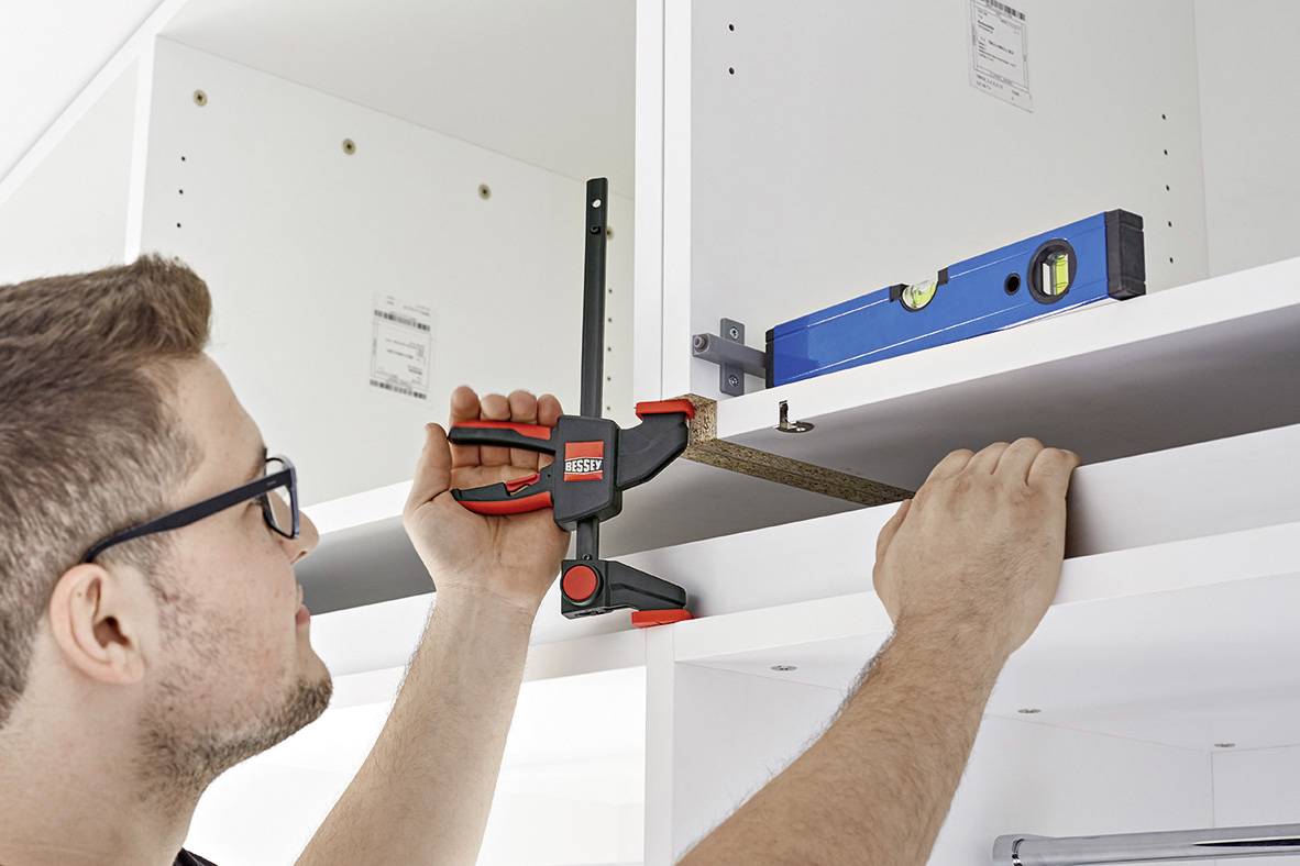 A man is securing a shelf board with a clamp. A spirit level is resting on the upper edge to align the shelf.