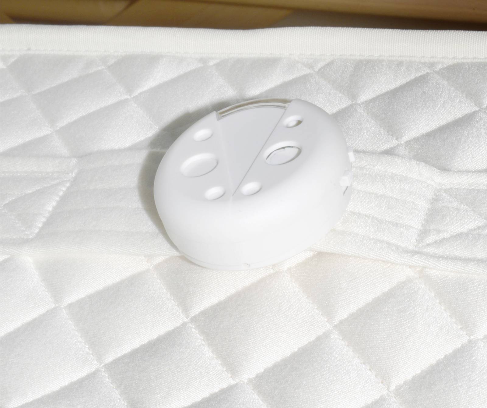 A white, round plastic button lies on a quilted mattress. The button has several holes and notches, possibly a fastener.