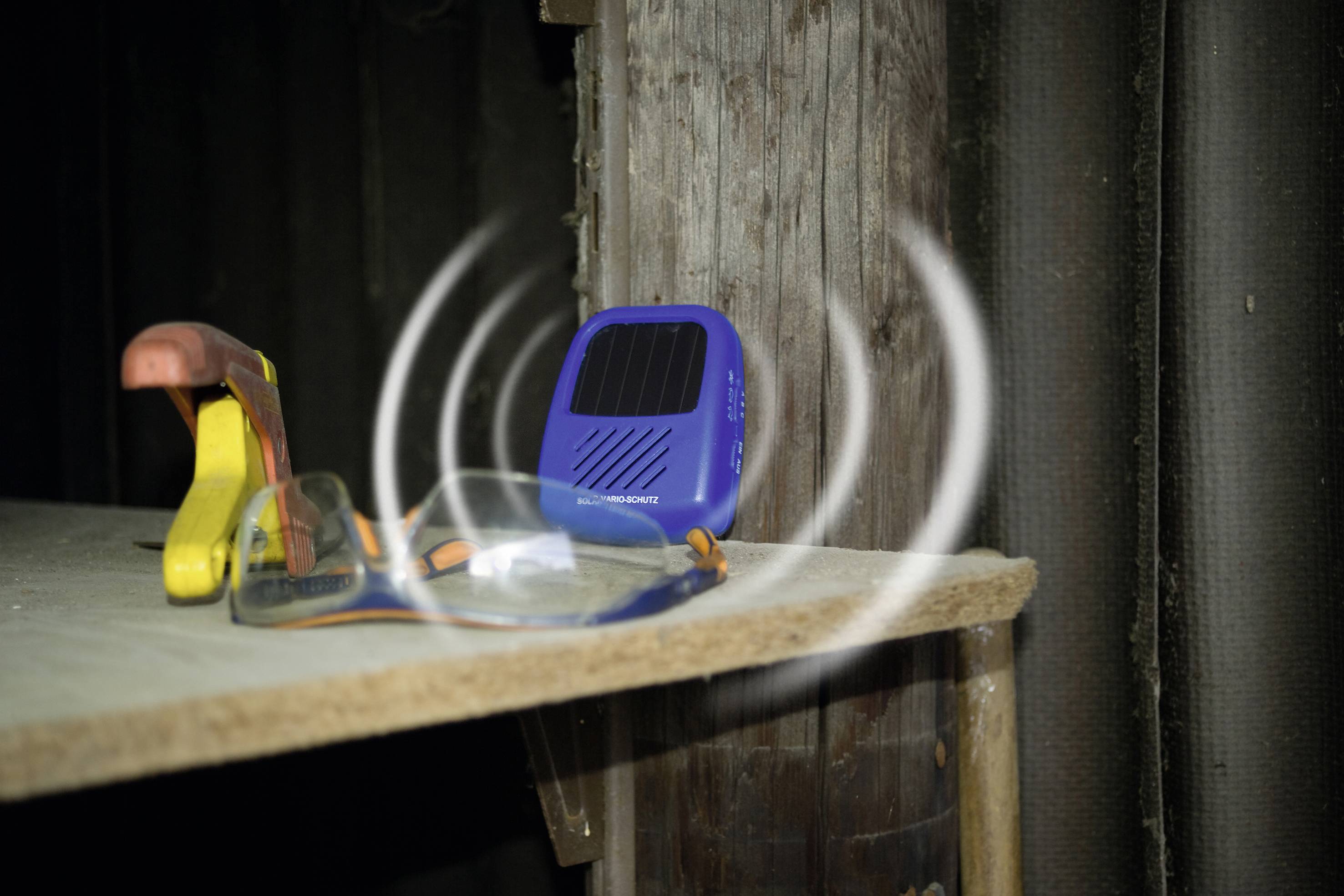 A blue device with a solar panel generates sound waves on a shelf next to safety glasses and a yellow tool.