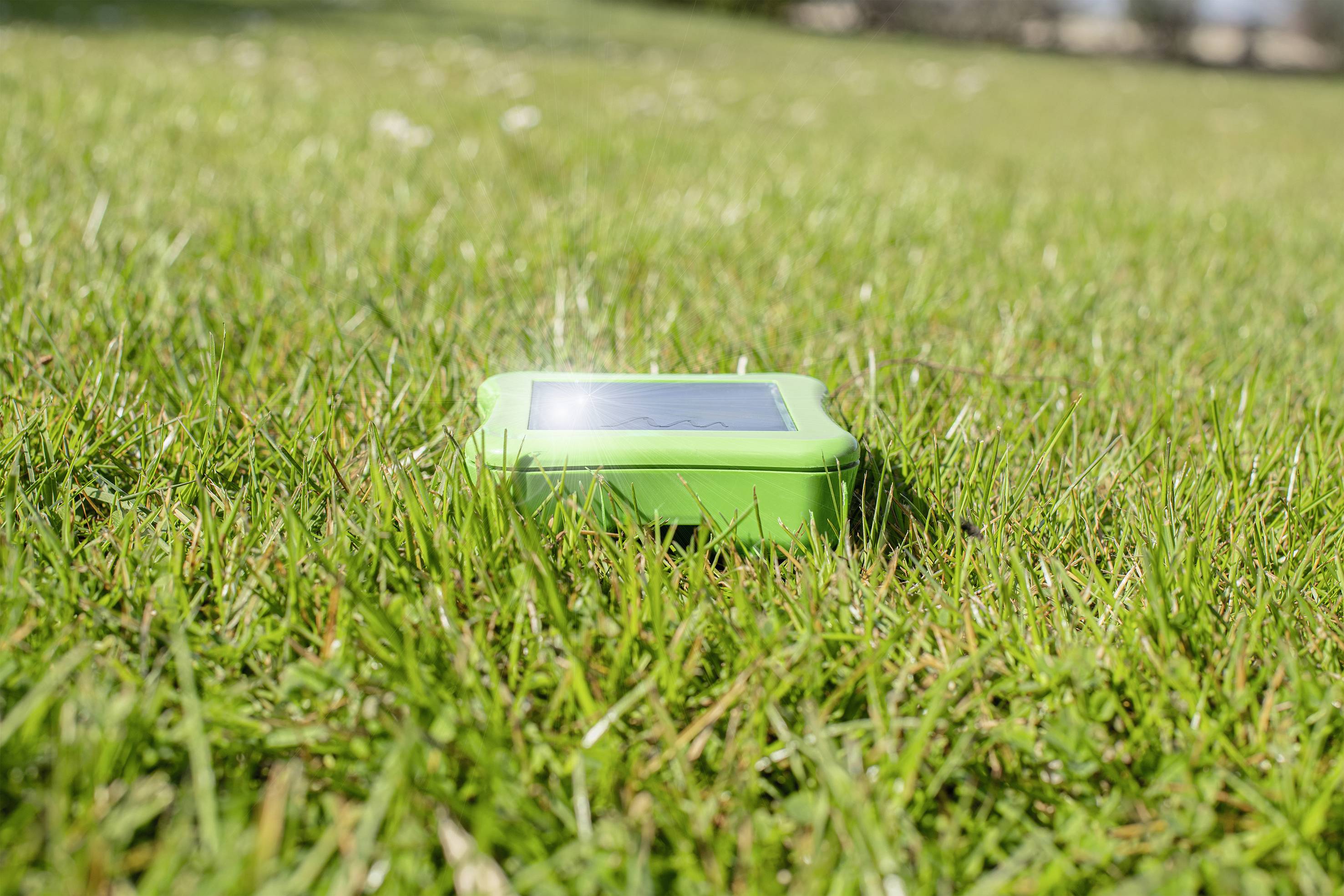 A green electronic device lies on a lawn, illuminated by sunlight, likely part of an automatic lawn sprinkler system.