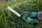 A person is trimming a hedge with a green electric hedge trimmer. The person is wearing work gloves.