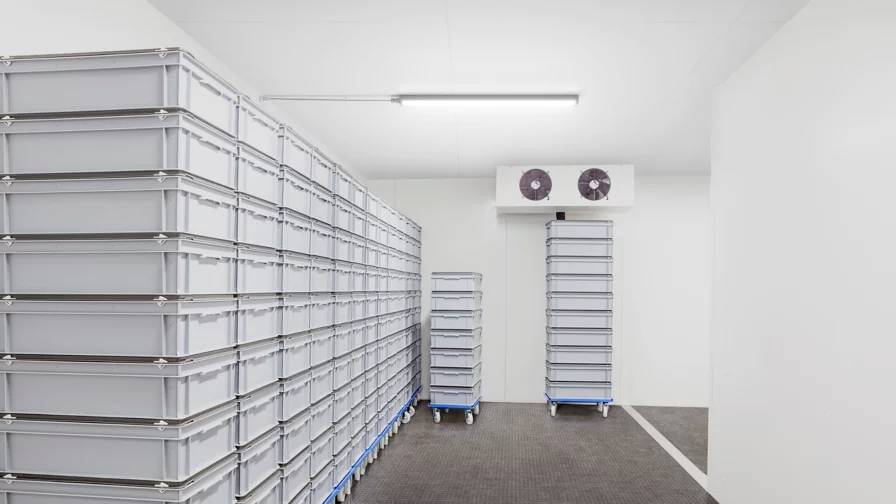 A storage room with stacks of large, gray plastic containers on movable carts, a cooling unit on the wall, and a bright overhead light.