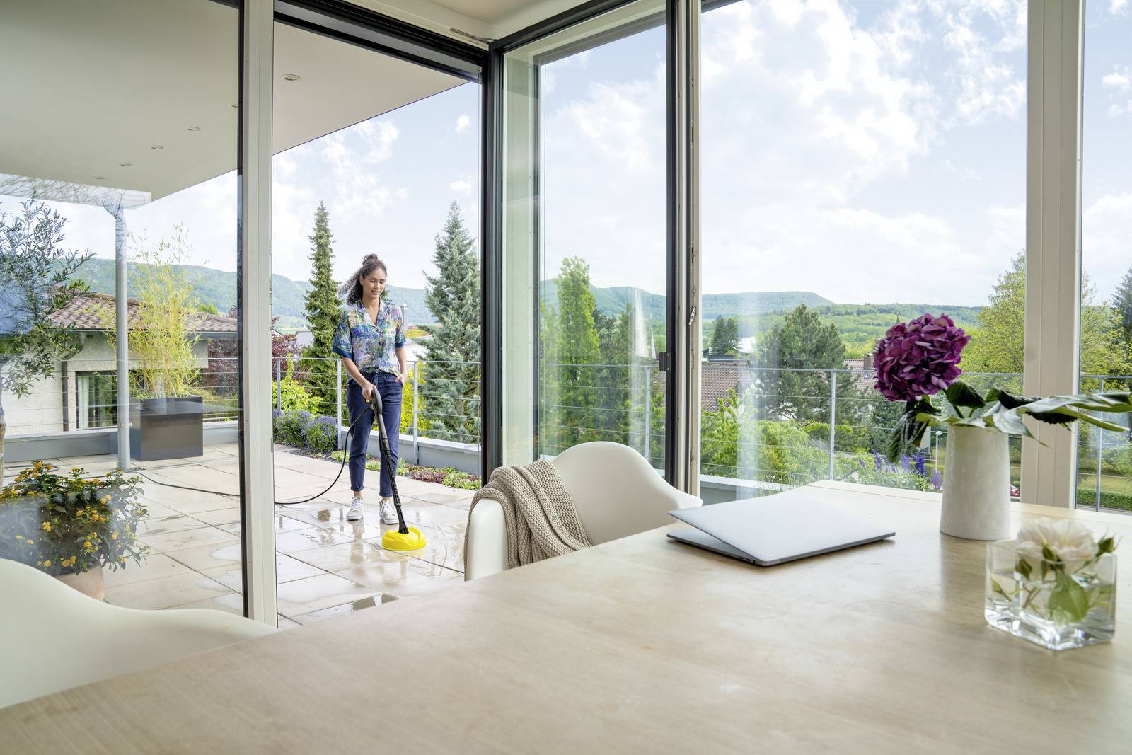 A woman uses a power cleaner on a patio outside, surrounded by greenery and mountains, while viewed through large glass doors from inside.