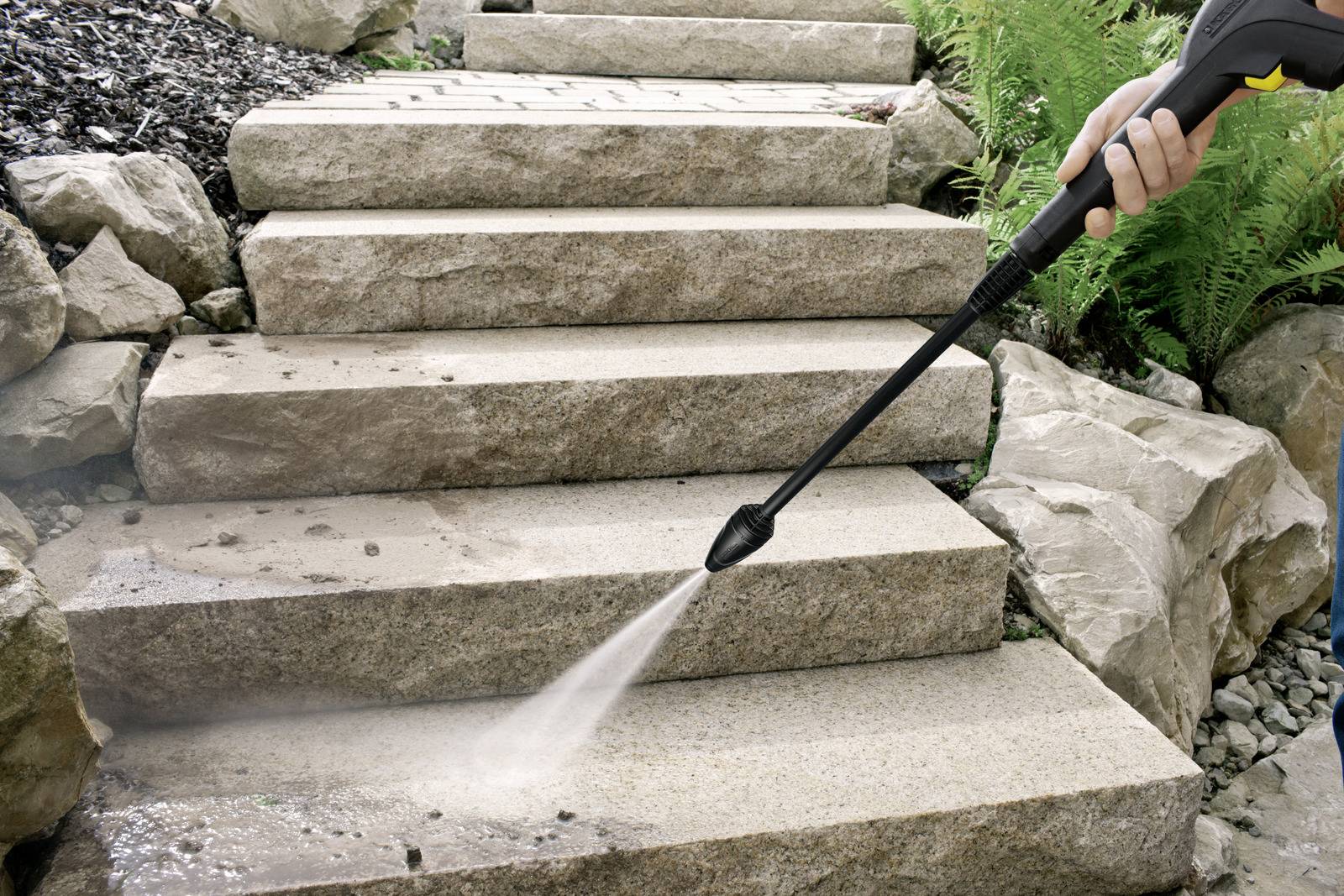 A person uses a pressure washer to clean stone steps surrounded by rocks and plants, showing effective outdoor cleaning.