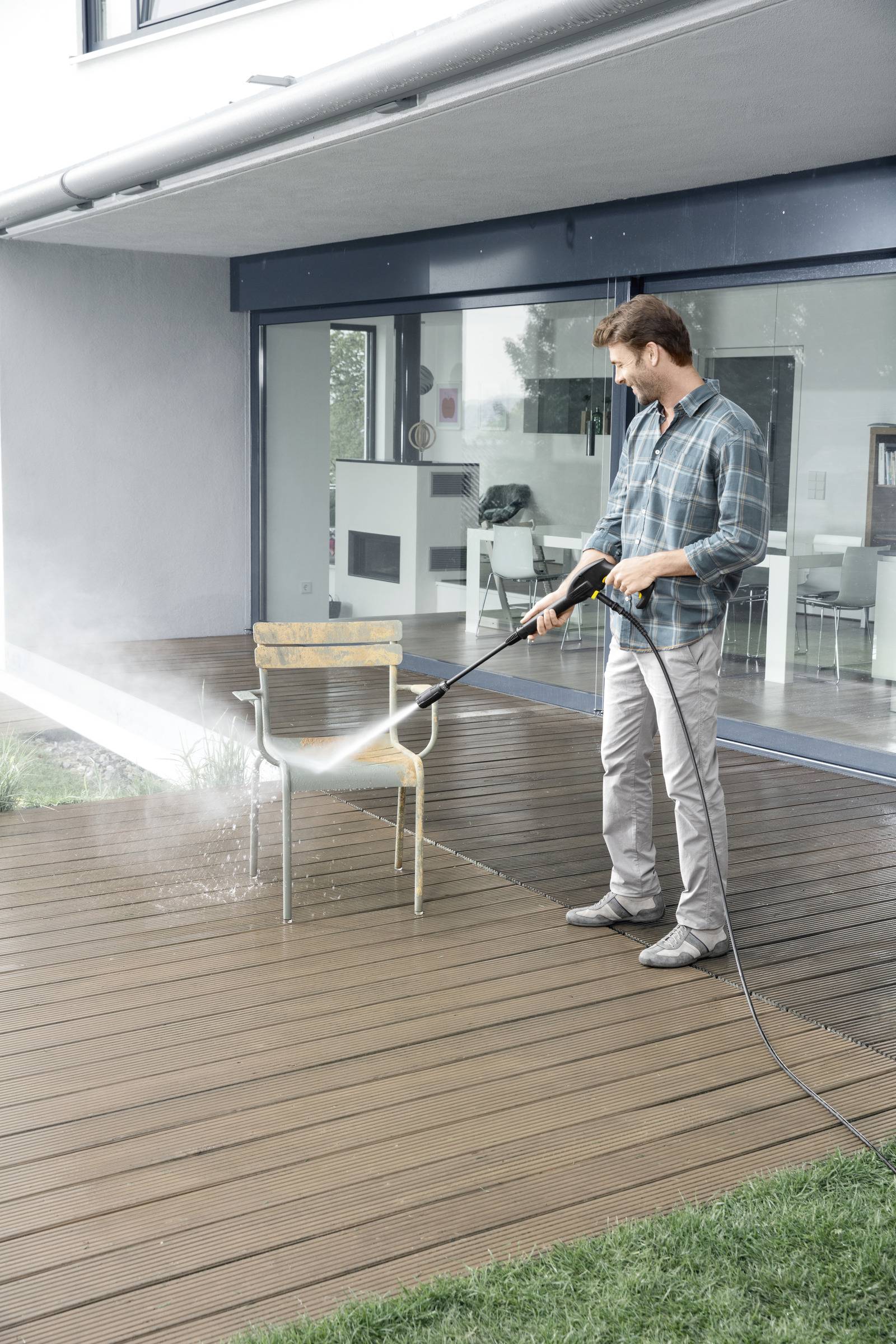 A person cleans a patio chair on a wooden deck using a pressure washer, with a modern house in the background.