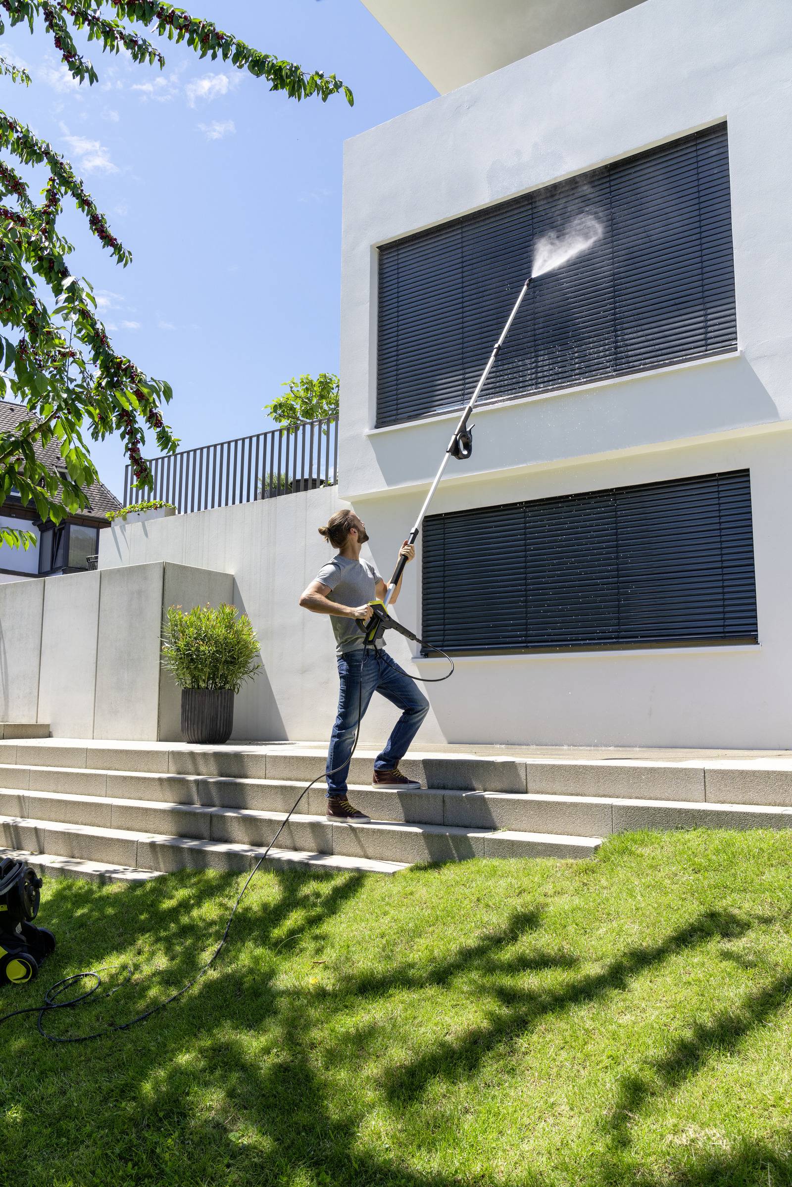 A person is cleaning the window shutters of a modern white house with a long pressure washer. Sunny day, green lawn.