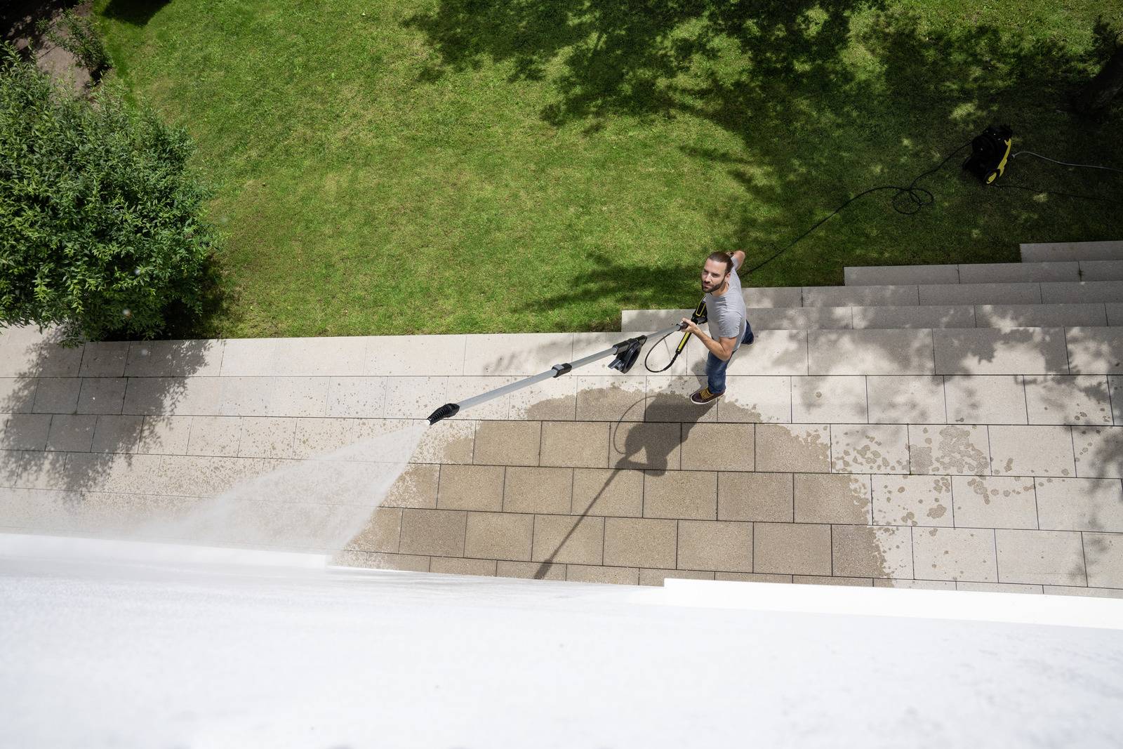A man is cleaning stone steps of an outdoor staircase with a pressure washer. Green grass can be seen in the background.