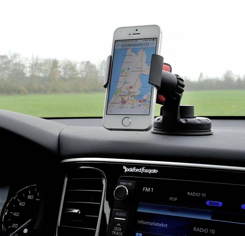 A smartphone displays a navigation map and sits in a car mount on the dashboard. Outside, a green landscape is visible.