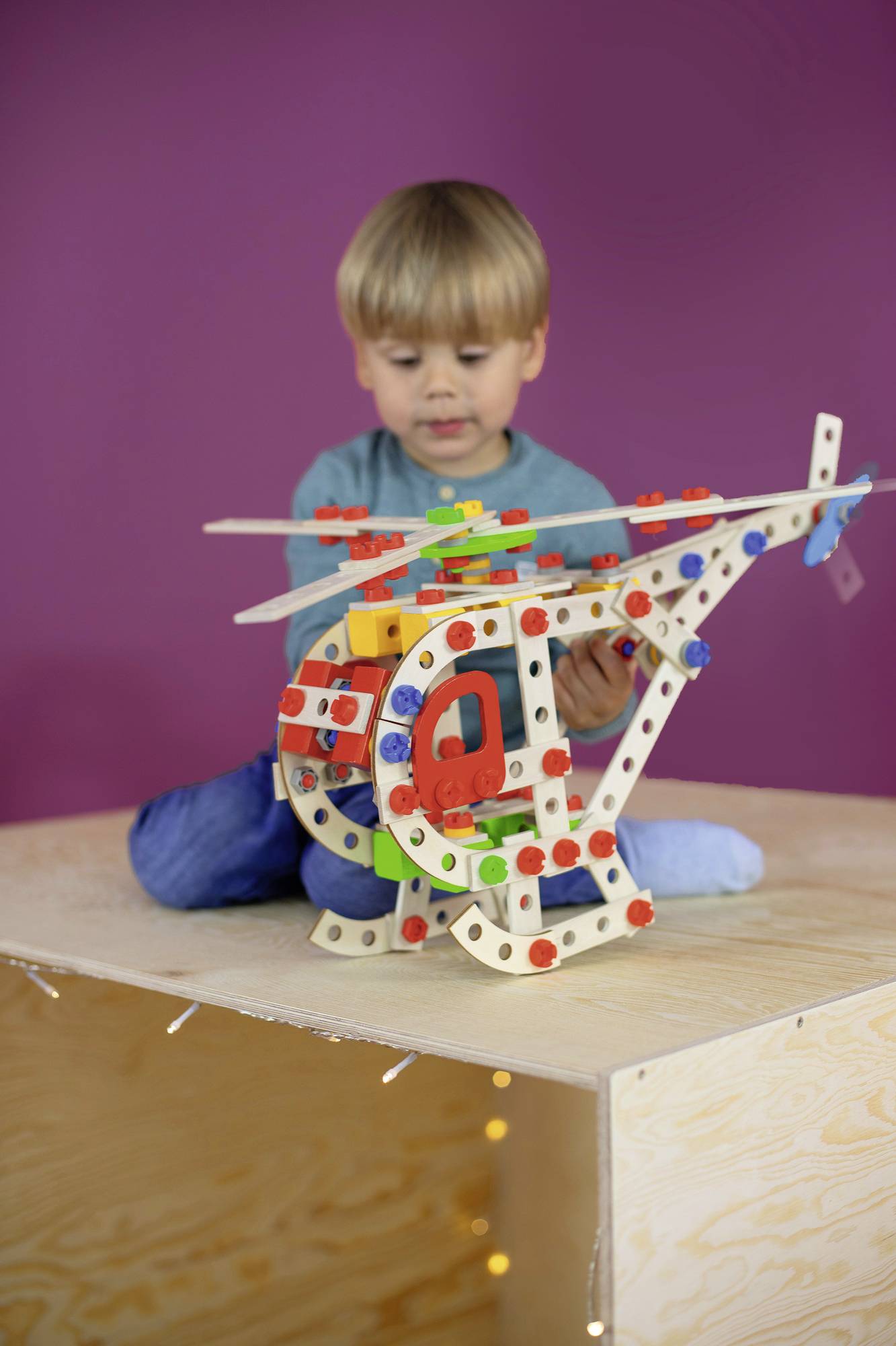 A child is playing with a colourful, modular helicopter on a wooden crate. The background is purple.