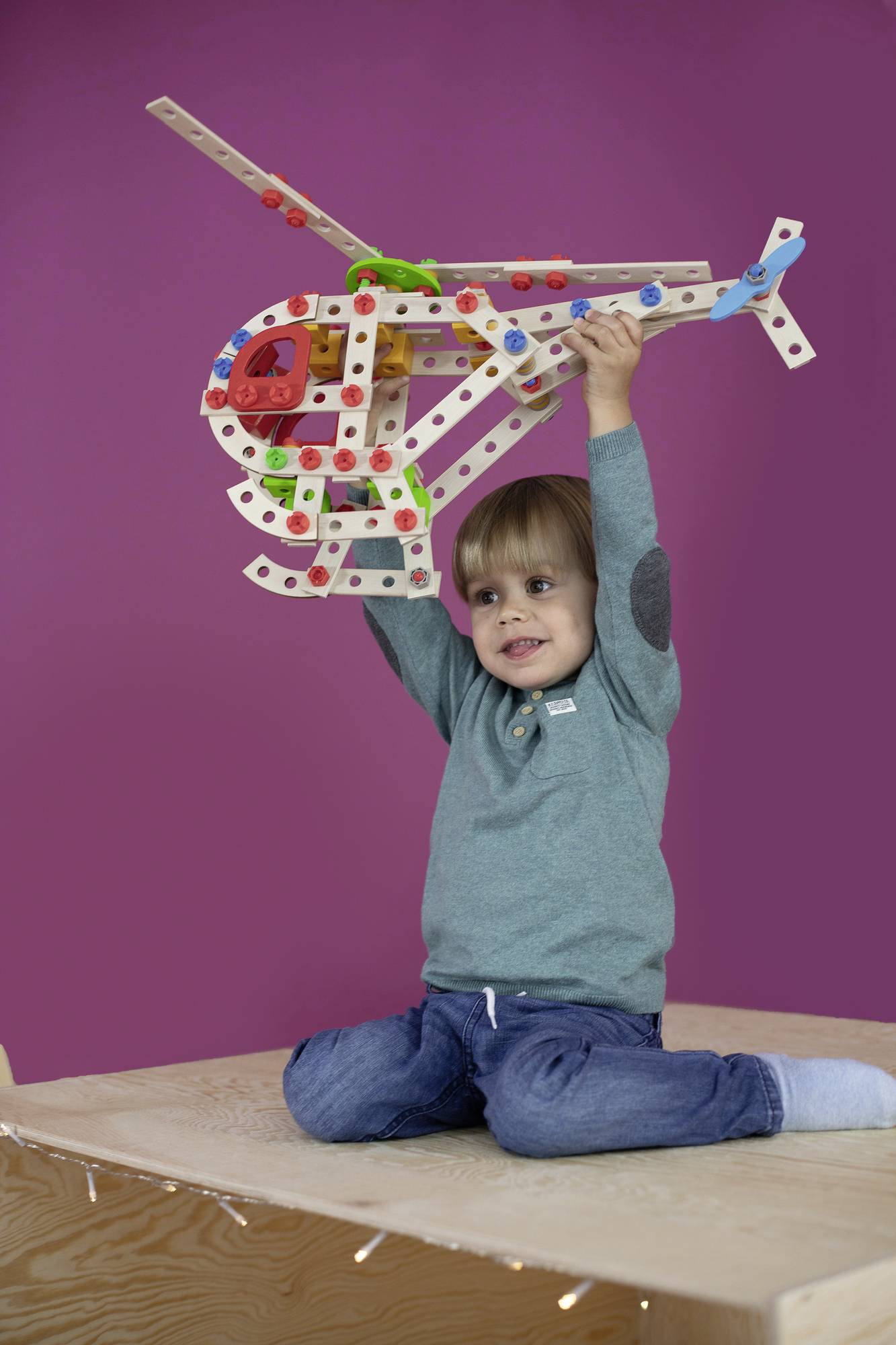 A child sits smiling on a table, holding a colourful, wooden model helicopter above their head.