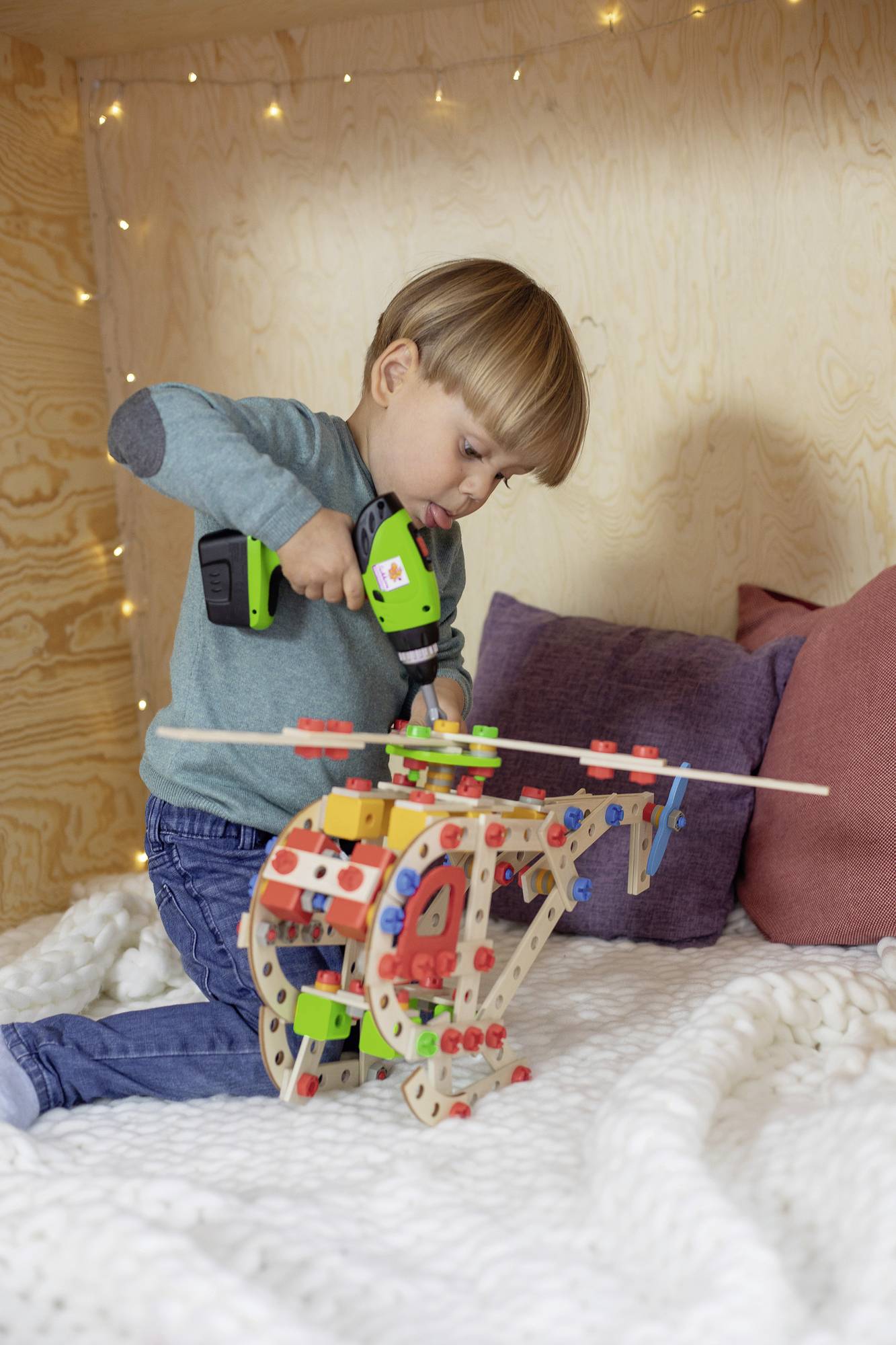 A child plays with a building set, using a toy drill to construct a colourful model. Bedroom with cosy lighting.