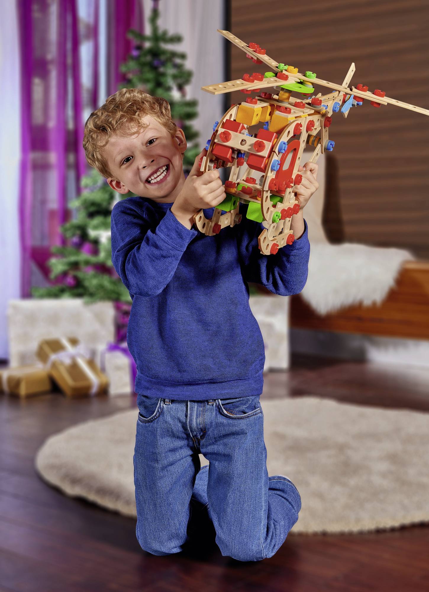 A boy is smiling and holding a colourful toy helicopter model in a living room. In the background, a decorated Christmas tree stands.